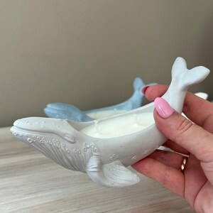 以下が含まれることがあります： 白いろうそくが入った灰色のクジラ型のキャンドルホルダー。ホルダーはセラミック製で、表面にテクスチャがあります。