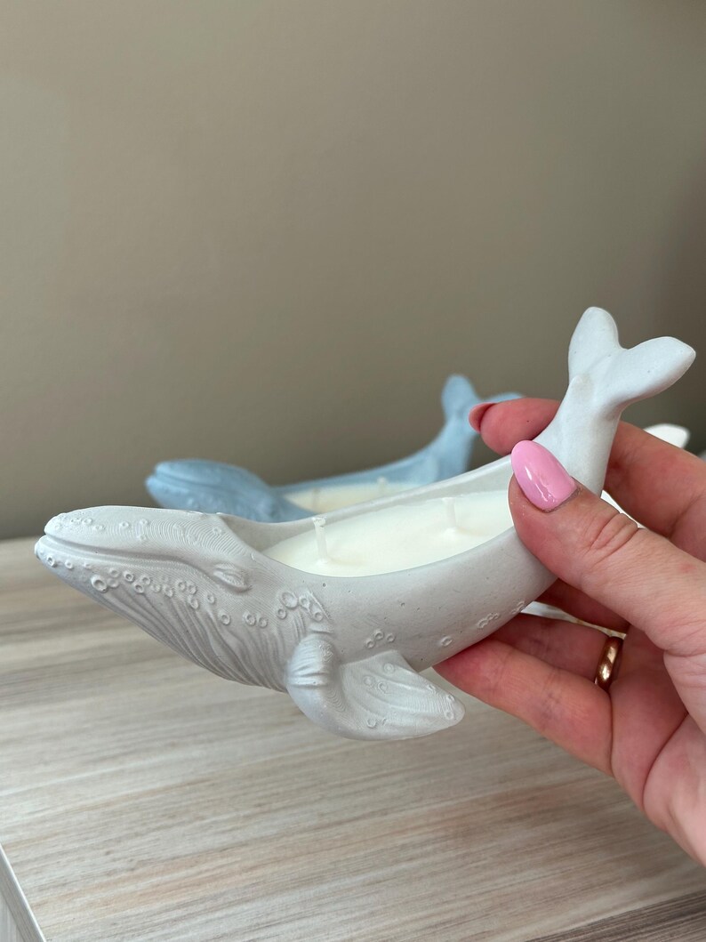 以下が含まれることがあります： 白いろうそくが入った灰色のクジラ型のキャンドルホルダー。ホルダーはセラミック製で、表面にテクスチャがあります。