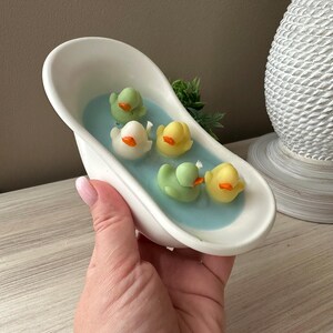 May include: A white ceramic bathtub-shaped container filled with light blue wax, featuring five small duck-shaped candles in shades of yellow and light green with orange beaks, arranged as if floating in the water within the miniature tub.