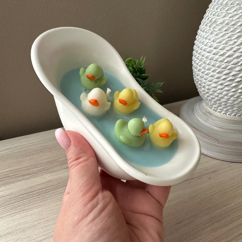 May include: A white ceramic bathtub-shaped container filled with light blue wax, featuring five small duck-shaped candles in shades of yellow and light green with orange beaks, arranged as if floating in the water within the miniature tub.