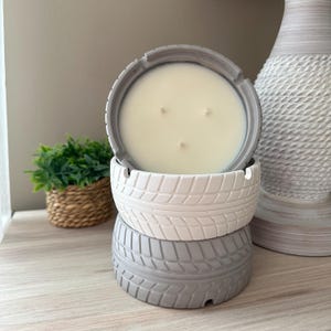 May include: A three-wick candle in a gray tire-shaped container. The candle is white and sits atop two additional tire-shaped containers, one white and one gray. The containers are stacked on top of each other. The candle is a decorative home accent.