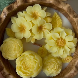 May include: A yellow floral candle in a wooden bowl. The candle features a variety of yellow flowers, including roses, peonies, and plumeria.