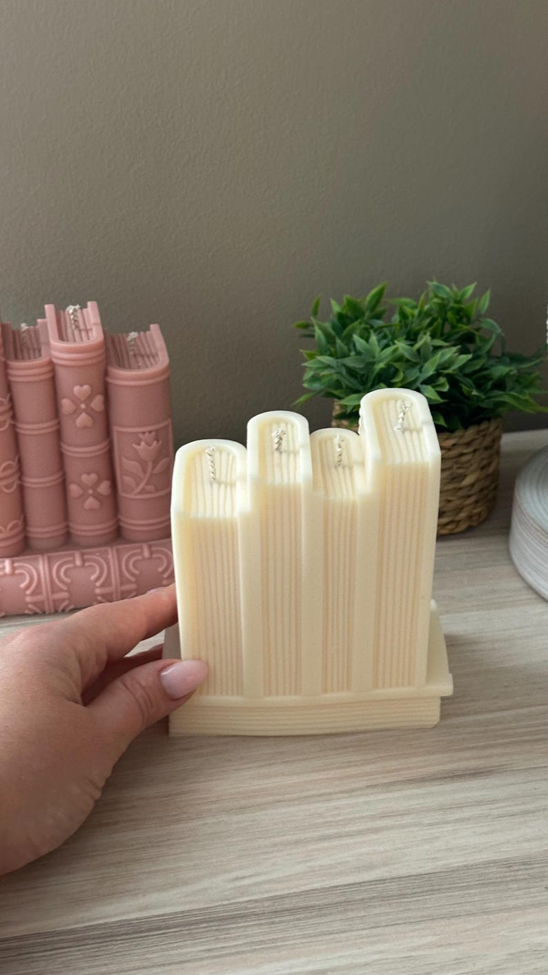 May include: Ivory-colored candle shaped like a stack of books, with vertical lines to mimic pages. The candle has four book-shaped sections with silver wicks. A pink candle shaped like books is in the background.