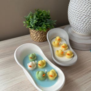 May include: Two oval-shaped bathtub-style candles. One white tub contains blue wax with four small duck-shaped wax figures in green, white, and yellow. The other gray tub contains white wax with four small yellow duck-shaped wax figures.