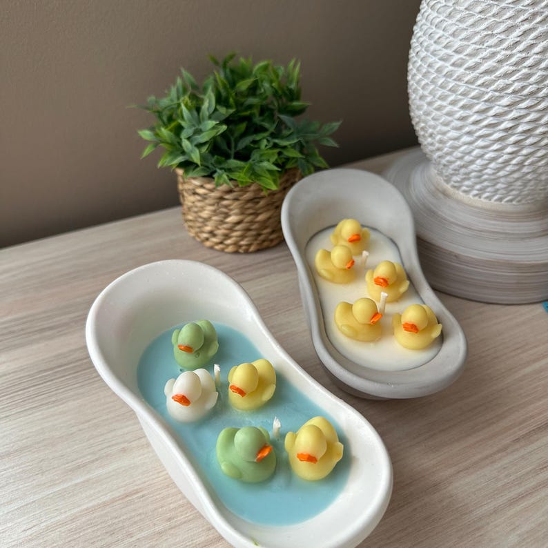 May include: Two oval-shaped bathtub-style candles. One white tub contains blue wax with four small duck-shaped wax figures in green, white, and yellow. The other gray tub contains white wax with four small yellow duck-shaped wax figures.