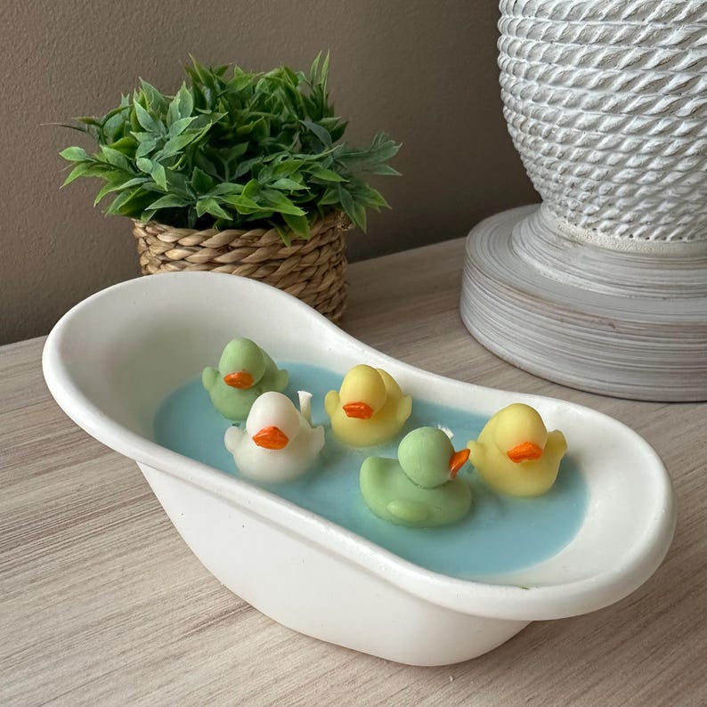 May include: A white ceramic bathtub-shaped container filled with light blue wax, featuring five small rubber duck-shaped wax figures in white, yellow, and light green with orange beaks. The duck figures are arranged across the blue wax surface.