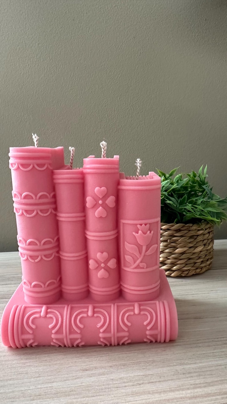 May include: A pink candle shaped like a stack of books. The candle has decorative details, including hearts and a flower. The wicks are visible on top. The candle is on a wooden surface.