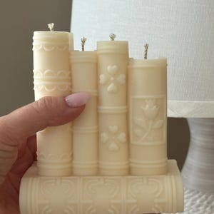 May include: Ivory-colored candle shaped like a stack of books. The candle features embossed designs of hearts, flowers, and decorative patterns. Each 'book' has a wick at the top, suggesting it's ready to be lit. A unique home decor item.