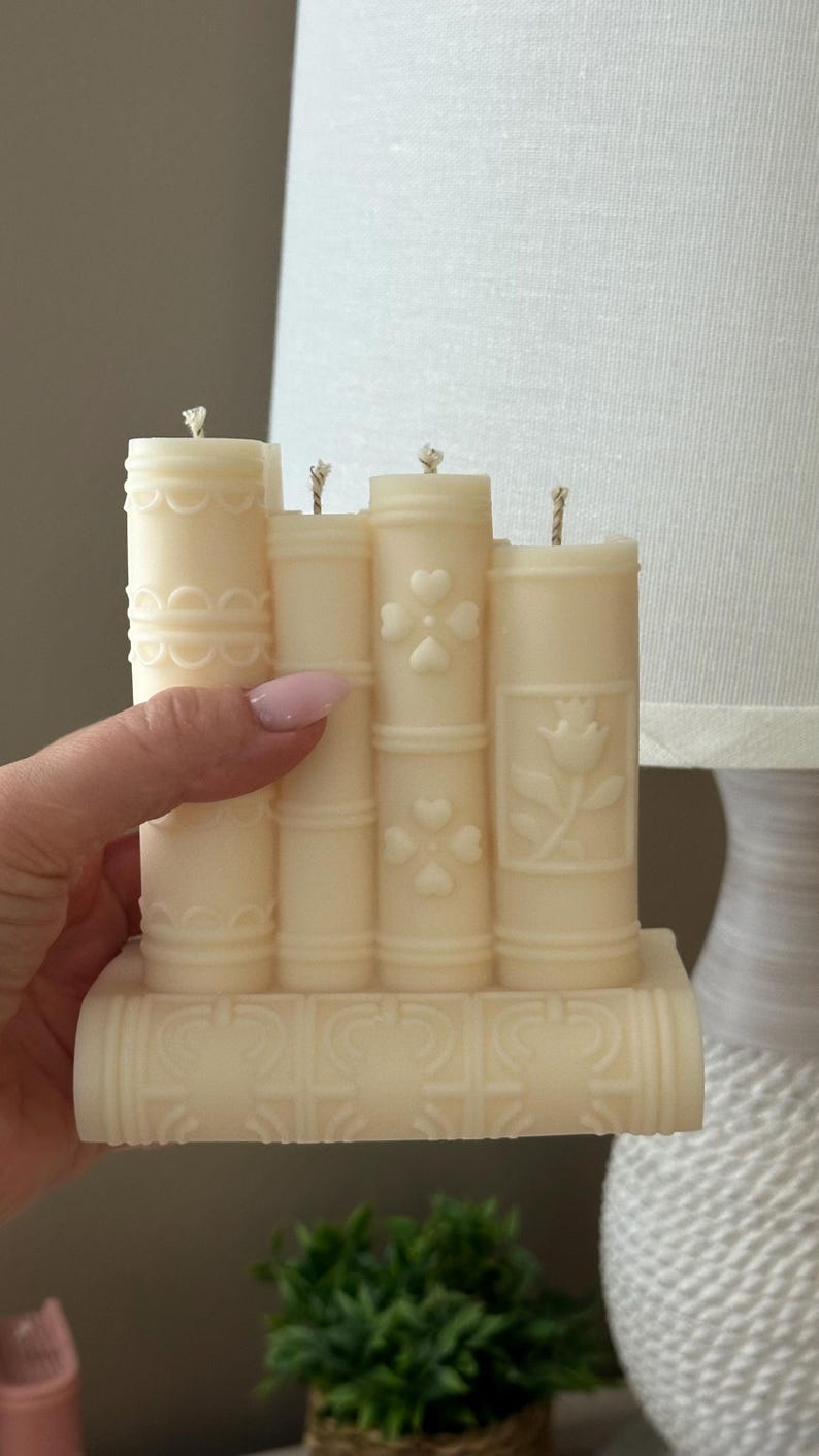 May include: Ivory-colored candle shaped like a stack of books. The candle features embossed designs of hearts, flowers, and decorative patterns. Each 'book' has a wick at the top, suggesting it's ready to be lit. A unique home decor item.