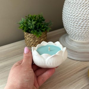 Op de afbeelding: Een witte keramische lotusbloemvormige kandelaar met een lichtblauwe kaars erin. De kaars heeft een enkele lont. De houder wordt door een persoon vastgehouden. Een kleine groene plant in een geweven mand staat op de achtergrond.