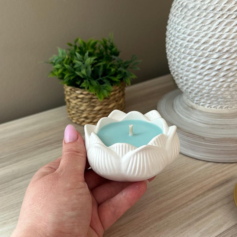 Op de afbeelding: Een witte keramische lotusbloemvormige kandelaar met een lichtblauwe kaars erin. De kaars heeft een enkele lont. De houder wordt door een persoon vastgehouden. Een kleine groene plant in een geweven mand staat op de achtergrond.