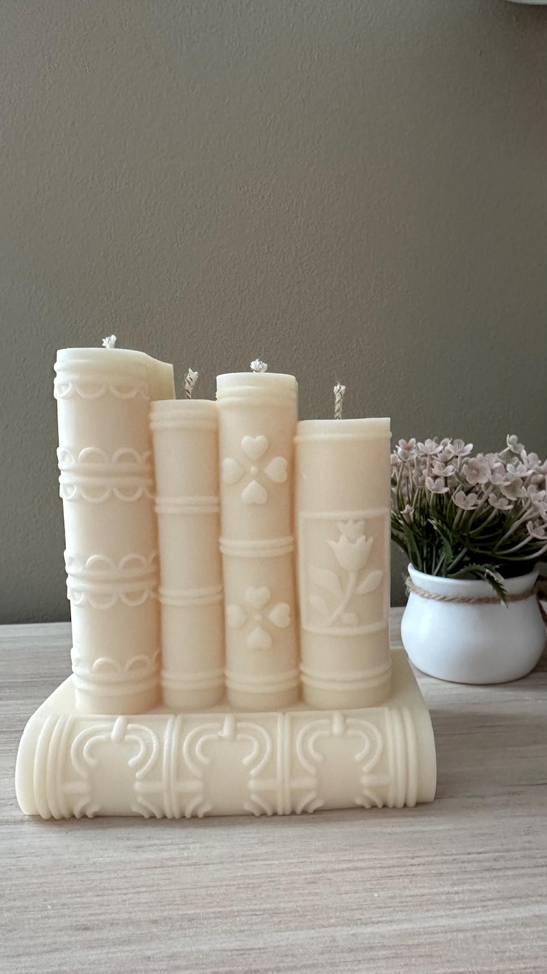 May include: Ivory-colored candle set shaped like a stack of books. Each candle has a different embossed design, including hearts and a tulip. The candles are on a base also shaped like a book, creating a unique decorative piece.