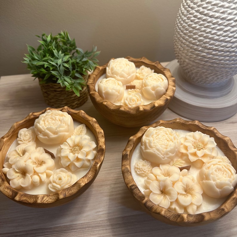 May include: Three wooden bowls filled with white floral scented candles. The candles are shaped like flowers and are arranged in a decorative pattern.