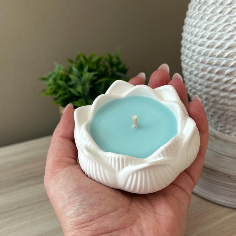Op de afbeelding: Een witte keramische kandelaar in de vorm van een lotusbloem, gevuld met lichtblauwe was. De kaars heeft een enkele lont. De houder wordt in een hand gehouden, met een kleine groene plant en een witte vaas met textuur op de achtergrond.