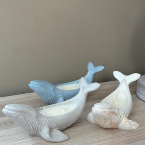 以下が含まれることがあります： 薄い青、薄いグレー、金色がかった白の3つのクジラ型のキャンドル。キャンドルは薄い木製の表面の上に置かれています。