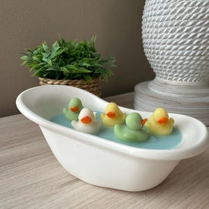 May include: A white ceramic bathtub-shaped container filled with light blue wax, featuring four small rubber duck-shaped wax figures in light green, white, and yellow colors, each with a bright orange beak, arranged in the blue wax surface.