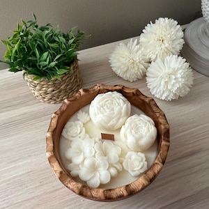 May include: A white floral scented candle in a wooden bowl. The candle is shaped like flowers and has a wooden wick. The bowl is made of natural wood and has a rustic look.