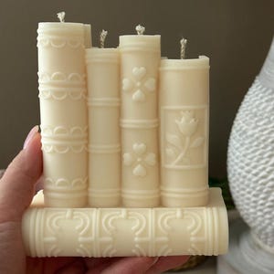 May include: Ivory-colored candle shaped like a stack of books. Each candle has a different embossed design, including hearts, flowers, and decorative patterns. The wicks are visible on top.