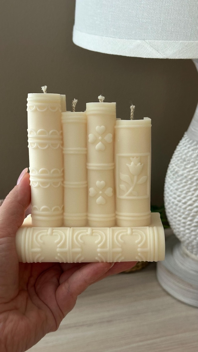 May include: Ivory-colored candle shaped like a stack of books. Each candle has a different embossed design, including hearts, flowers, and decorative patterns. The wicks are visible on top.