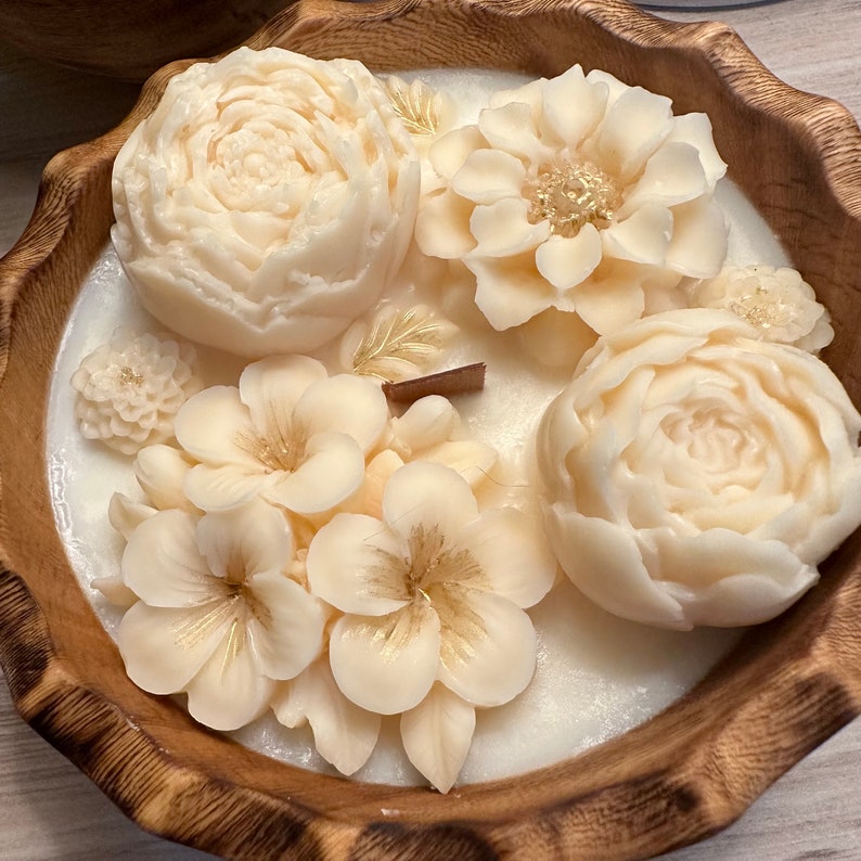 May include: A white candle in a wooden bowl with a floral design. The candle is decorated with white flowers and gold accents.