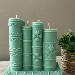 May include: A set of four teal-colored candles shaped like books. The candles are decorated with embossed designs, including hearts and a floral pattern. The candles are on a base that resembles a stack of books.