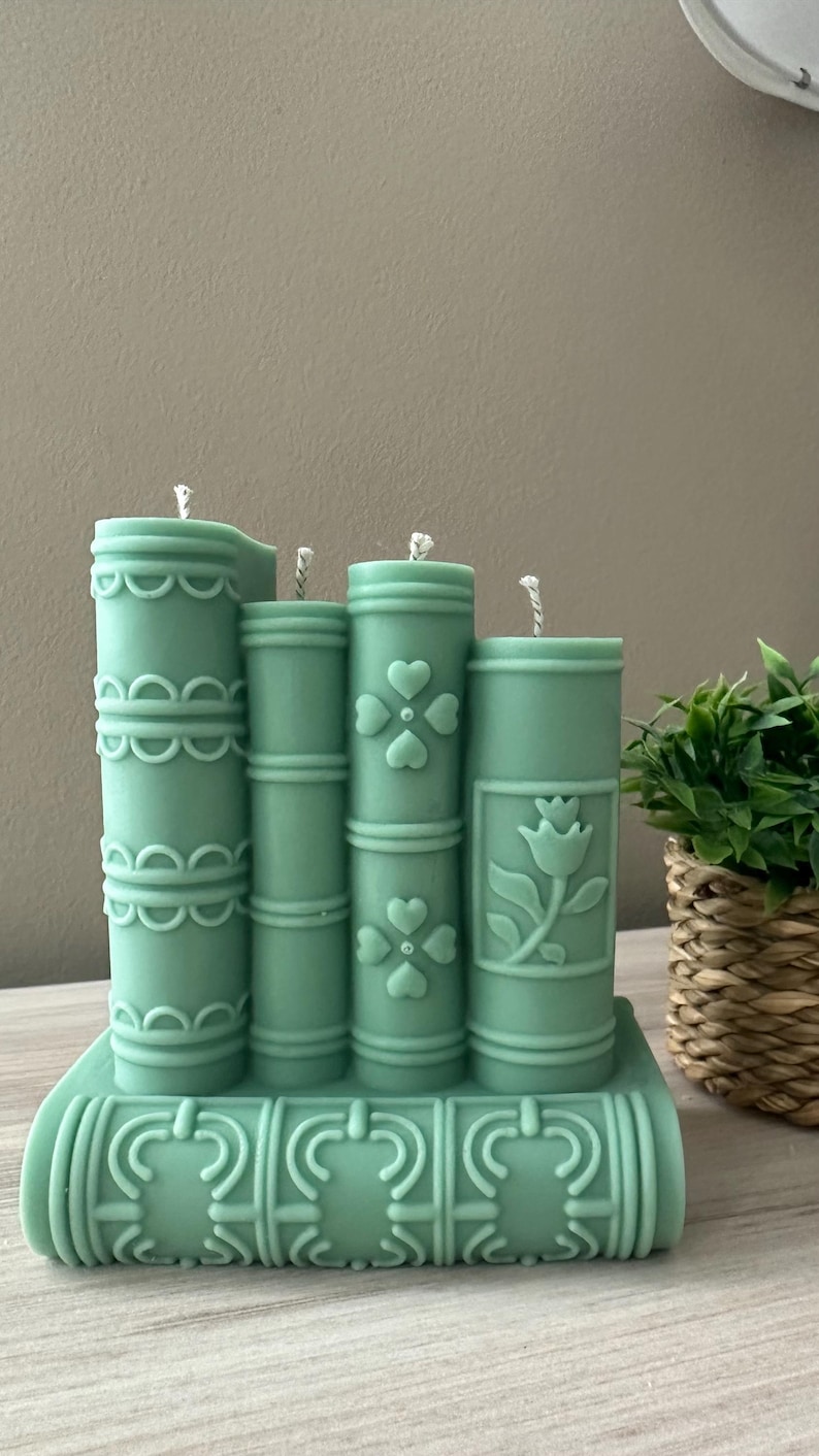 May include: A set of four teal-colored candles shaped like books. The candles are decorated with embossed designs, including hearts and a floral pattern. The candles are on a base that resembles a stack of books.