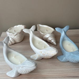 以下が含まれることがあります： 貝殻とクジラの形をした装飾的なろうそくが4本。ろうそくは白、水色、金色です。ろうそくは薄い茶色の木製の表面の上に置かれています。