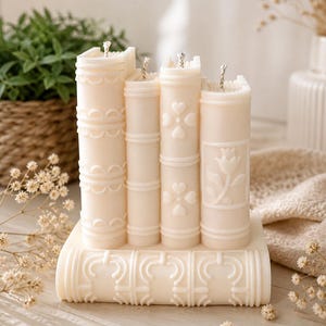 May include: A set of ivory-colored candles shaped like stacked books. Each candle has a unique embossed design, including hearts and floral patterns. The candles are arranged on a base with decorative detailing, creating a literary-themed decorative piece.