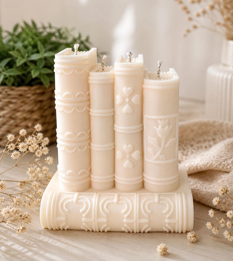 May include: A set of ivory-colored candles shaped like stacked books. Each candle has a unique embossed design, including hearts and floral patterns. The candles are arranged on a base with decorative detailing, creating a literary-themed decorative piece.