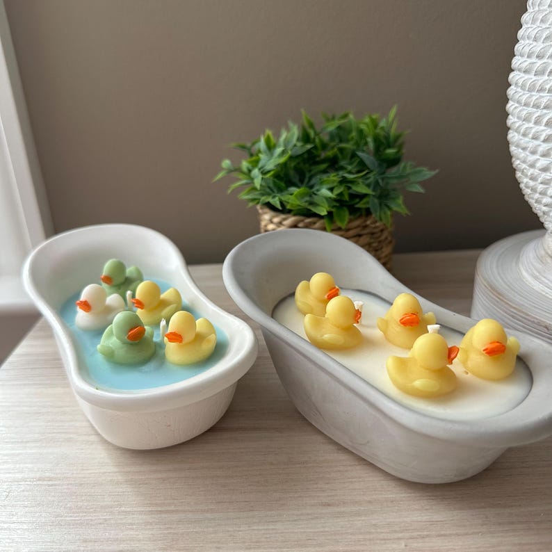 May include: Two white ceramic bathtub-shaped containers filled with wax. The left tub contains blue wax with two light green and two yellow duck-shaped wax pieces. The right tub contains white wax with four yellow duck-shaped wax pieces.