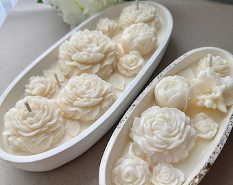 May include: Two ivory-colored, oval-shaped candles with floral designs. Each candle features detailed, carved roses and other flower shapes. The candles are set in white containers, with a white flower arrangement in the background.