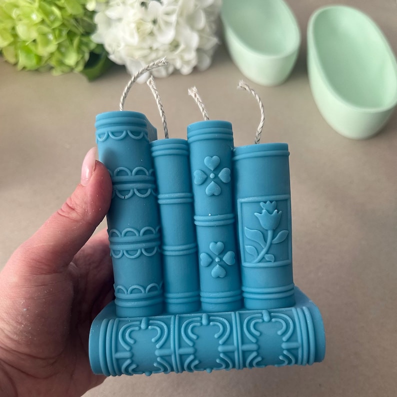 May include: A teal-colored candle shaped like a stack of books. The candle features decorative details, including floral and geometric patterns. Each book has a wick. The candle is held by a hand, with flowers and other objects in the background.