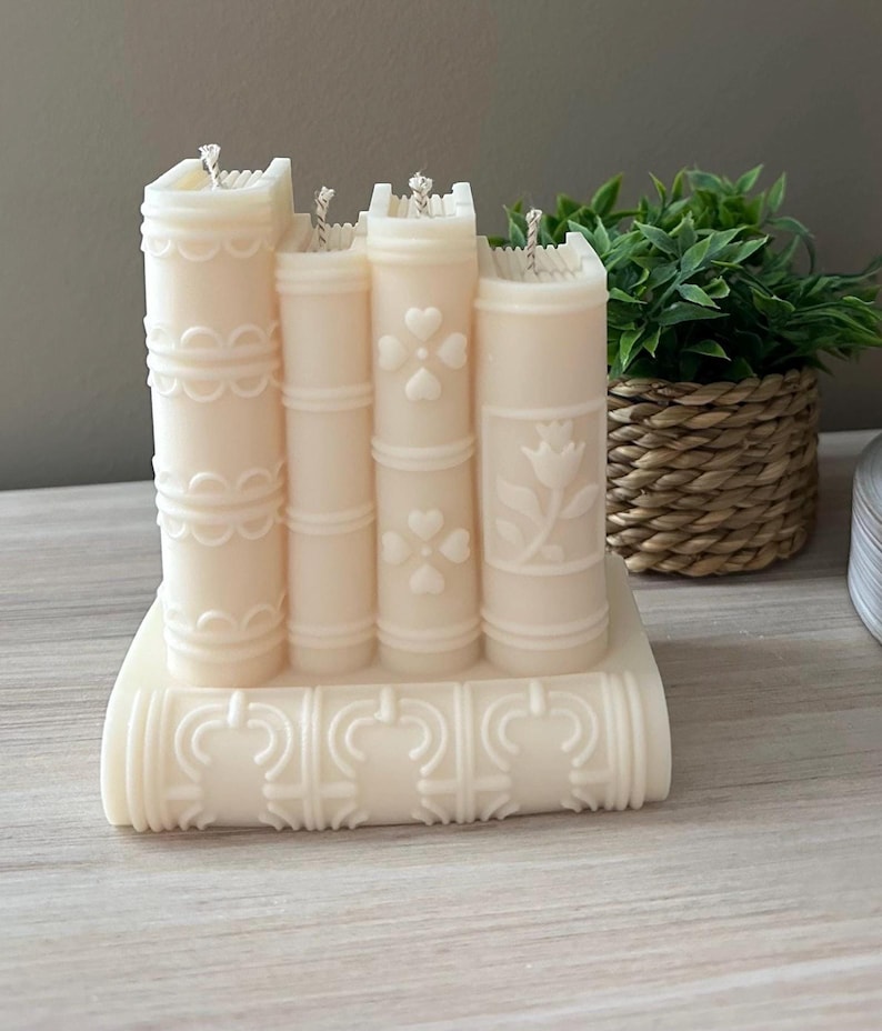 May include: Ivory-colored candle shaped like a stack of books. The candle features detailed designs, including hearts, floral patterns, and decorative edges. The wicks are visible on top. A decorative item for home decor.