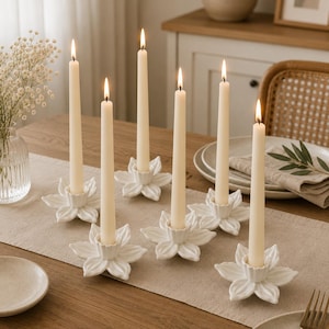 May include: A collection of lit, ivory-colored taper candles in white ceramic flower-shaped candle holders. The candles are arranged on a beige table runner, with a vase of white flowers and a wooden table in the background.