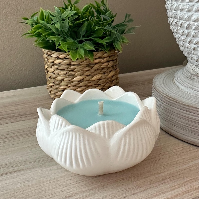 Op de afbeelding: Een witte keramische lotusbloemvormige kandelaar met een lichtblauwe kaars en een enkele lont. Een kleine groene plant in een geweven mand en een decoratieve witte lampvoet staan op de achtergrond.