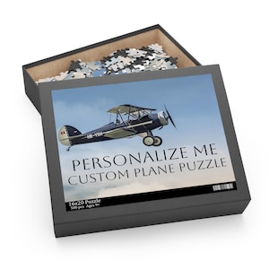 May include: A 500-piece jigsaw puzzle featuring a black and white biplane flying in a blue sky. The puzzle box has the text "Personalize Me Custom Plane Puzzle" and "16x20 Puzzle 500 pcs Ages 9+".