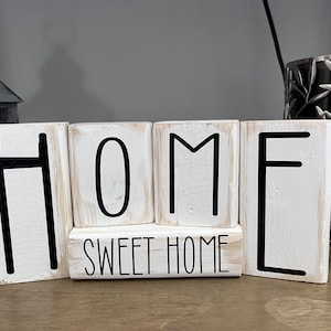 Peut inclure: Blocs de bois blancs orthographiant "HOME" avec un bloc plus petit en dessous indiquant "SWEET HOME".