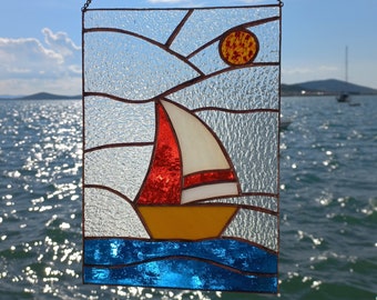 May include: A stained glass window hanging with a sailboat and a sun in a blue sky. The sailboat is red, white, and yellow. The sun is orange and yellow.