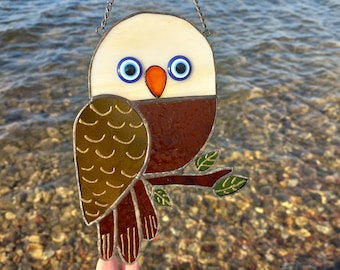 May include: A stained glass owl with brown, gold, and green accents. The owl has large blue eyes with white pupils and an orange beak. The owl is hanging from a silver chain.