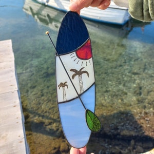 Può includere: Una tavola da surf in vetro colorato con un design blu, bianco e rosso. La tavola da surf ha due palme e un sole su di essa. C'è una pagaia verde attaccata alla tavola da surf.