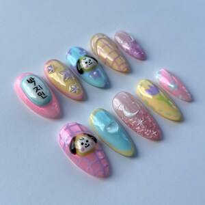 May include: Set of ten pastel-colored fake nails with various designs. The designs include a cartoon dog, stars, a crescent moon, and a checkered pattern. One nail has the Korean text "방탄소년단" written on it.