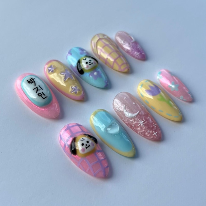May include: Set of ten pastel-colored fake nails with various designs. The designs include a cartoon dog, stars, a crescent moon, and a checkered pattern. One nail has the Korean text "방탄소년단" written on it.