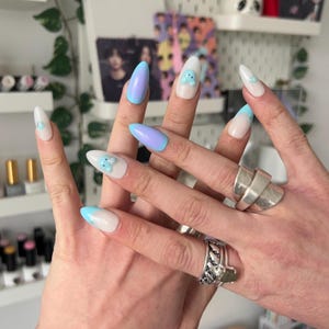 May include: A hand displays long, almond-shaped nails. Some nails are painted with a gradient of light blue to lavender, while others feature a white base with light blue tips or a small blue bear design. Silver rings adorn the fingers.