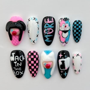 May include: Set of ten false nails with various designs.  The designs include a checkered pattern, a cartoon character, a cartoon face, and text that says "Jack in the Box" and "More".