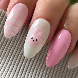 Puede incluir: Primer plano de una mano con u&ntilde;as en forma de almendra de color rosa y blanco. Una u&ntilde;a presenta un conejito rosa con ojos negros.