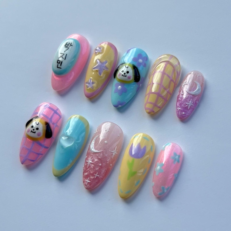 May include: Set of 10 pastel-colored fake nails with various designs, including a cartoon dog, stars, a crescent moon, and a flower. One nail has the Korean text "빵지민".