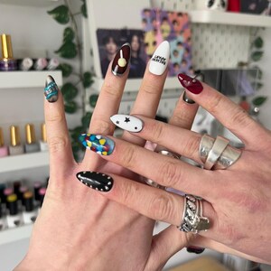 May include: A close-up of hands showcasing a variety of nail art designs. The nails feature diverse styles, including a black nail with silver studs, a white nail with black stars, and a nail with colorful abstract shapes. Other nails have a brown and silver design, a white nail with text, and a deep red nail.