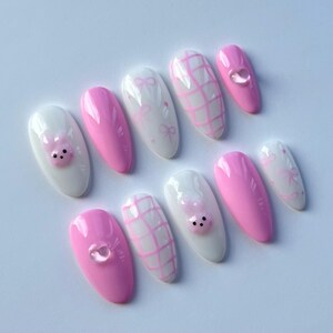 Puede incluir: Conjunto de diez u&ntilde;as postizas ovaladas de color rosa y blanco con varios dise&ntilde;os. Algunas u&ntilde;as tienen una carita de cerdo rosa, otras tienen un patr&oacute;n de cuadros blancos y otras tienen un patr&oacute;n de lazo blanco.