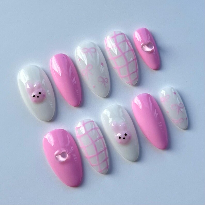 Puede incluir: Conjunto de diez u&ntilde;as postizas ovaladas de color rosa y blanco con varios dise&ntilde;os. Algunas u&ntilde;as tienen una carita de cerdo rosa, otras tienen un patr&oacute;n de cuadros blancos y otras tienen un patr&oacute;n de lazo blanco.