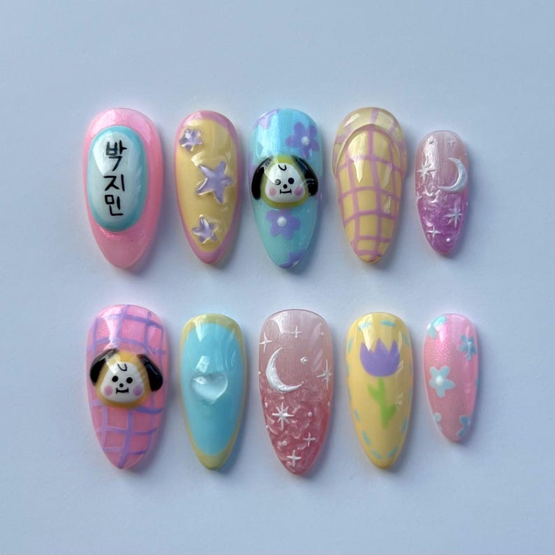 May include: Set of ten pastel-colored fake nails with various designs. The designs include a cartoon dog, stars, flowers, a crescent moon, and a checkered pattern. One nail has the Korean text "박지민" written on it.